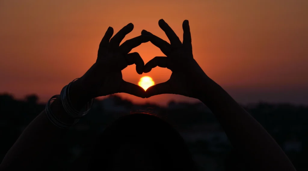Person's hands making heart in front of sunset